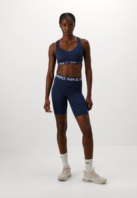 Sutien de sport bleumarin și pantaloni scurți de ciclism, cu accente contrastante ale logo-ului Nike alb. Țesătură netedă, design ajustat, stil minimalist, asociat cu adidași ușori.
