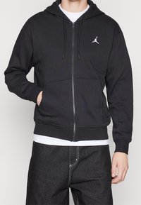 Persona che indossa una felpa nera con zip e logo Jumpman bianco, una maglietta bianca sotto e jeans scuri con cuciture bianche visibili.