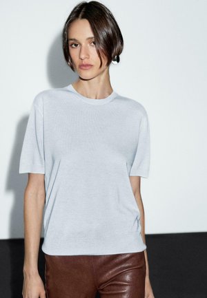 Model stands against a plain backdrop, wearing a light gray short-sleeve shirt and brown leather pants, with a neutral expression.
