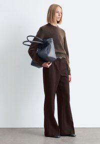 Frau in braunem Pullover und weit geschnittenen Hosen, die eine große schwarze Leder-Handtasche hält und gegen eine weiße Wand steht.