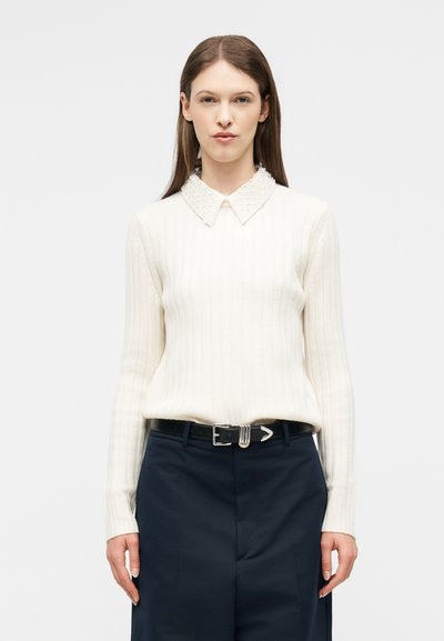 Femme aux cheveux bruns raides portant un pull crème côtelé avec un col perlé et un pantalon noir taille haute avec ceinture noire, debout devant un fond blanc.