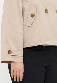 Beige trenchjacka med dubbelknäppt design, stora bruna knappar och manschettdetalj. Slätt tyg, kortare längd och skräddarsydd passform.