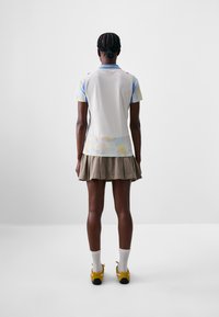 Polo à manches courtes avec un motif abstrait pastel, présentant un dos beige, des accents bleus et une jupe plissée kaki. Chaussettes blanches et chaussures jaunes.