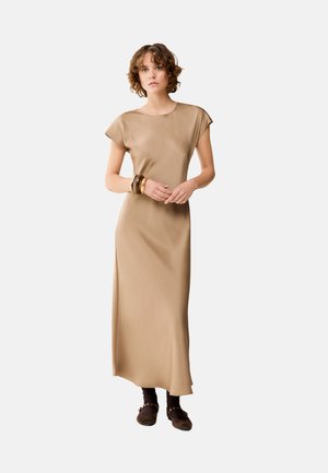 Vrouw met kort krullend haar, gekleed in een lange beige jurk met kapmouwen, bruine armbanden, donkere sokken en bruine schoenen, staande met gevouwen handen.