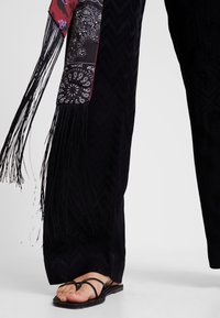 Pantalon noir avec des détails en franges, comportant un panneau en tissu à motifs. Accompagné de sandales noires à brides, mettant en valeur les orteils.