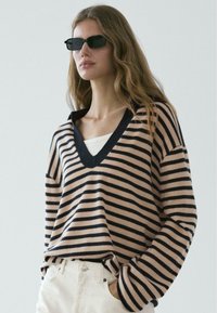 Maglione a righe beige e blu navy, con scollo a V e maniche lunghe. Il materiale sembra morbido, con una vestibilità comoda. Abbinato a pantaloni bianchi.