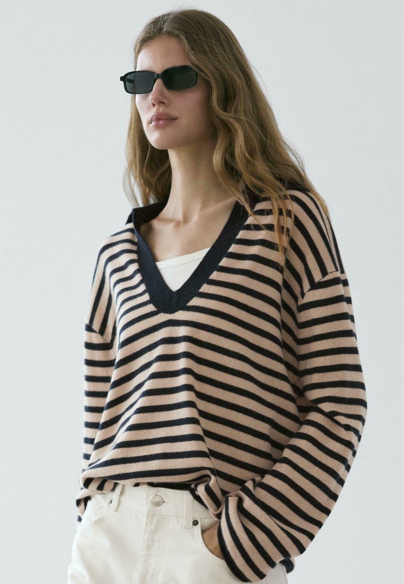 Maglione a righe beige e blu navy, con scollo a V e maniche lunghe. Il materiale sembra morbido, con una vestibilità comoda. Abbinato a pantaloni bianchi.