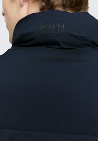Zadný pohľad na osobu oblečenú v tmavomodrej bunde s vypnutým golierom, na ktorom je značka "DENIM TOM TAILOR".