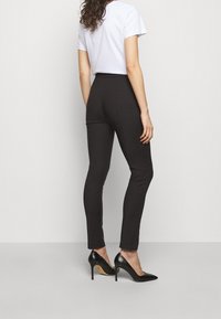 Schwarze, enganliegende Hose aus glattem Stoff, hoch geschnitten, kombiniert mit einem weißen T-Shirt und schwarzen High Heels, von hinten betrachtet.