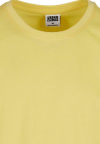 Camiseta amarilla de cuello redondo hecha de tela suave; la etiqueta dice "URBAN CLASSICS", talla M, con costuras visibles a lo largo del escote.