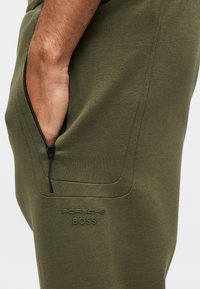 Χέρι στην τσέπη με φερμουάρ πράσινου ελιάς παντελονιού με το λογότυπο «Porsche BOSS» ανάγλυφο κάτω από την τσέπη.