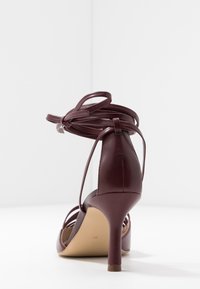 Sandal a tacco alto bordeaux in pelle liscia con cinturini incrociati e dettaglio alla caviglia legato; presenta un sottile tacco a spillo.