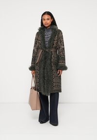 Μακρύ παλτό με σχέδια σε σκούρο πράσινο χρώμα, με ψεύτικη γούνα και ζώνη. Διαθέτει περίπλοκα σχέδια. Συνδυάζεται με ανοιχτό καφέ τσάντα και φαρδιά τζιν.