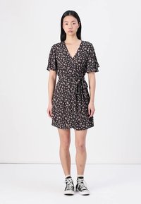 Robe portefeuille noire à motifs floraux avec de courtes manches volantes, taille à nouer et longueur au-dessus du genou, associée à des baskets montantes noires.