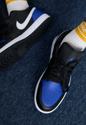 Stopy w czarnych, niebieskich i białych butach Nike Air Jordan, z czarnymi sznurowadłami i białych skarpetkach z żółtymi paskami na niebieskim dywanie.