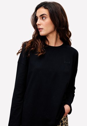 Junge Frau mit langen, welligen Haaren, die einen schwarzen Sweatshirt und eine Hose mit Leopardenmuster trägt, schaut nach links und hat eine Hand in ihrer Tasche.