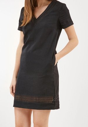 Femme portant une robe noire courte avec manches courtes, col en V, poches latérales, et un détail décoratif à ourlet perforé.