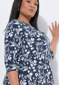 Top blu navy con motivo floreale e maniche 3/4. Il materiale è morbido, caratterizzato da fiori bianchi e foglie a contrasto. Scollo tondo.