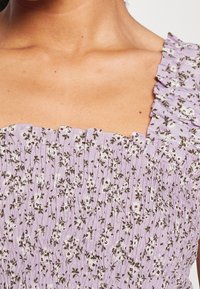 Fialové květinové šaty s nařaseným designem, s čtvercovým výstřihem a volánkovými okraji. Materiál zahrnuje malé bílé a zelené květy.