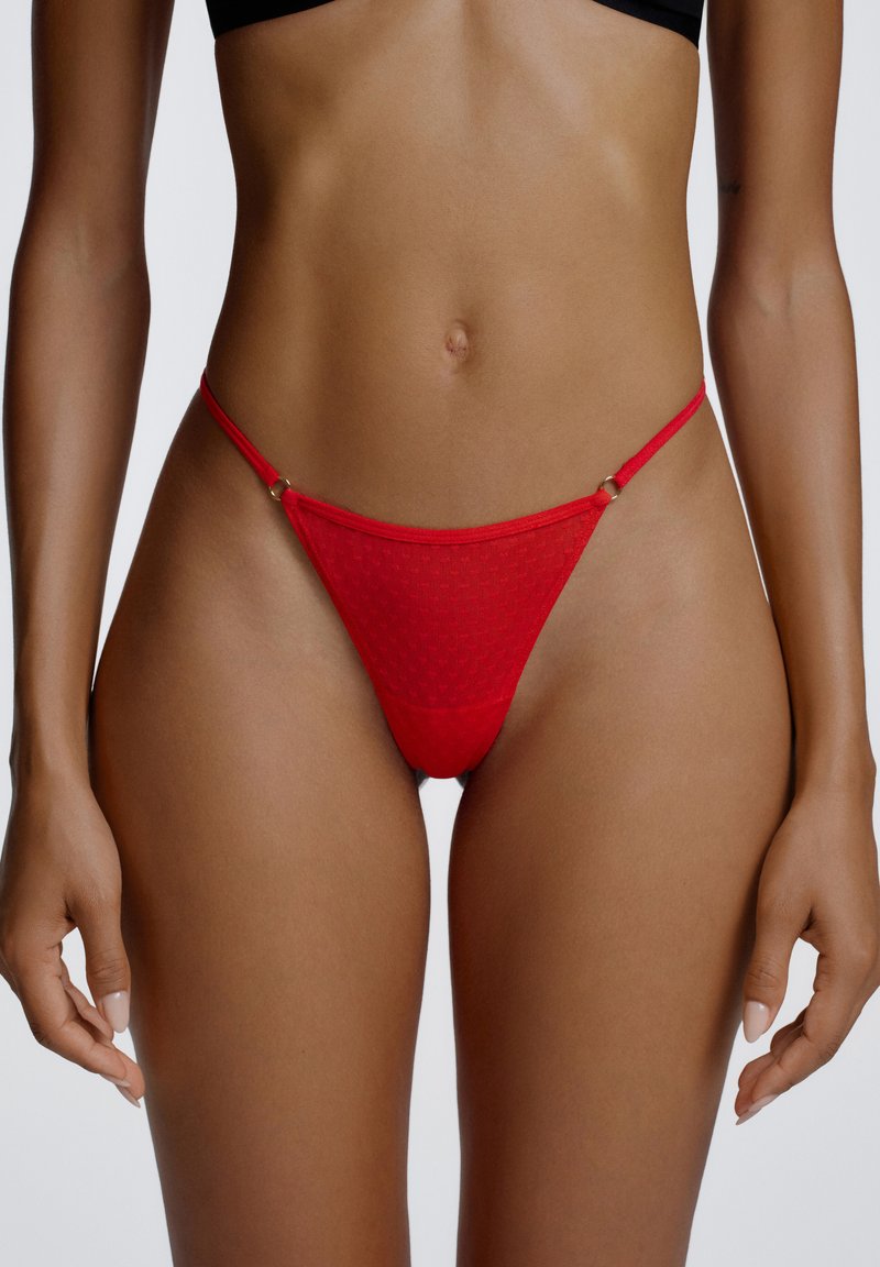 String porte-jarretelles en maille rouge avec fines bretelles, doté d'une surface texturée et de petits accessoires métalliques dorés sur les côtés.