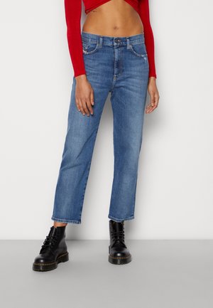 Nainen, joka käyttää lyhennettyjä sinisiä farkkuja, mustia nauhallisia saappaita ja punaista pitkähihainen lyhyt paita, seisoo yksiväristä taustaa vasten.