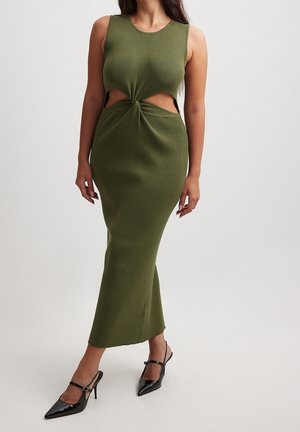 Femme portant une robe en maille vert olive sans manches avec des découpes latérales et des chaussures à talons hauts noires à bouts pointus, debout devant un fond uni.