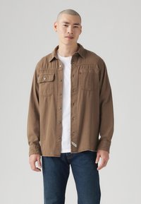 Camisa marrón de botones con dos bolsillos en el pecho, costuras visibles y un corte relajado, llevada sobre una camiseta blanca, combinada con unos jeans azules.
