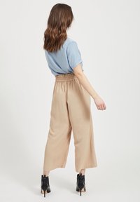 Jasnoniebieska bluzka z krótkim rękawem w połączeniu z beżowymi spodniami o szerokich nogawkach, z elastycznym pasem. Modelka nosi czarne botki na obcasie.