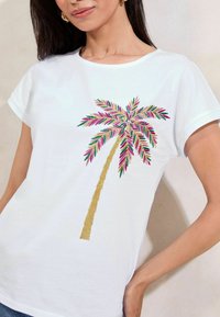 T-shirt en coton blanc avec une broderie de palmier colorée aux accents roses, verts et dorés. Manches courtes et coupe décontractée.
