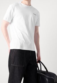 Uomo che indossa una t-shirt bianca di Calvin Klein e pantaloni neri, mentre tiene una borsa di pelle nera per i manici su uno sfondo bianco.