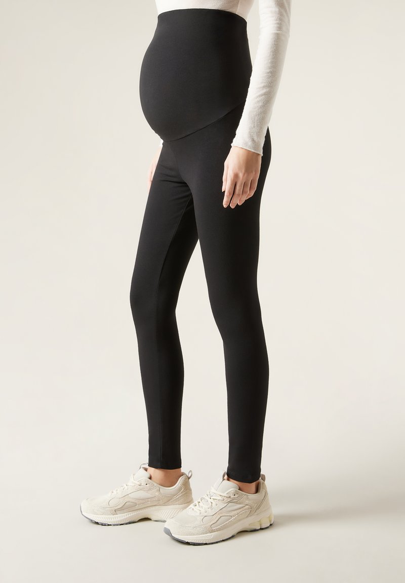 Schwarze Schwangerschaftsleggings mit einem hohen, dehnbaren Bund. Weicher, glatter Stoff mit enger Passform, kombiniert mit hellen Sportschuhen.