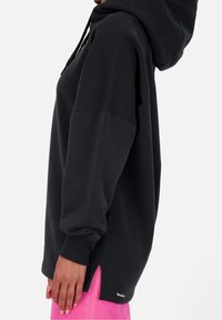 Felpa nera con cappuccio, maniche lunghe, spacchi laterali e polsini a coste. Il tessuto sembra morbido. Indossata sopra pantaloni rosa, con un dettaglio del logo all'orlo.