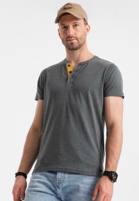 Šedé tričko s krátkým rukávem typu henley se žlutým zapínáním, dvěma knoflíky a měkkou texturou. Nosit s světle modrými džínami a béžovou kšiltovkou.