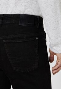 Black denim jeans with a smooth texture, featuring a leather patch at the waistband, angled back pockets, and subtle stitching details.