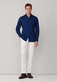 Camicia blu navy con bottoni, realizzata in tessuto liscio, caratterizzata da maniche lunghe e orlo stondato, abbinata a pantaloni bianchi e scarpe marroni.