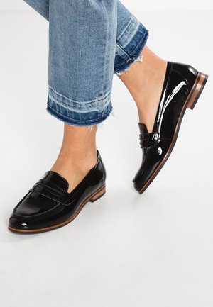 Mocassins en cuir verni noir avec semelles en bois, portés avec un jean bleu à ourlet effiloché et retroussé sur des pieds nus, sur fond blanc.