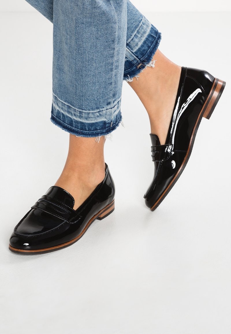 Mocassins en cuir verni noir avec semelles en bois, portés avec un jean bleu à ourlet effiloché et retroussé sur des pieds nus, sur fond blanc.
