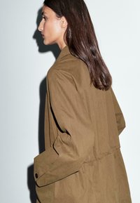Brun trenchcoat med en afslappet pasform, lavet af let stof. Har en indsnævret talje, knapærmer og et simpelt design. Sideprofil vist.