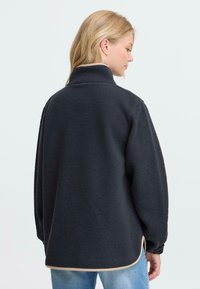 Svart fleece-pullover med høy krage, løs passform og teksturert stoff. Merkbar kontrastsøm ved kanten og sideventiler.