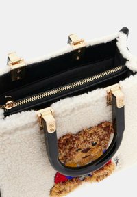 Valkoinen huopakassi, jossa on mustat keinonahkakahvat. Kullanväriset yksityiskohdat ja vetoketjusuljenta. Toisella puolella on brodeerattu karhudesign.