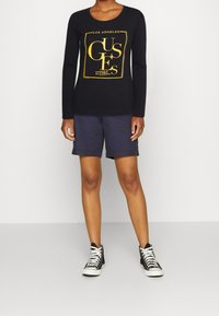 Osoba v černém tričku s dlouhým rukávem s žlutým logem "GUESS Los Angeles Est. 1981", námořnických šortkách a černých sneakers s vysokým střihem a bílými tkaničkami.