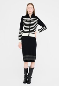 Donna che indossa un maglione zip-up a righe bianche e nere e una gonna nera abbinata, con stivali neri fino a metà polpaccio, in piedi davanti a uno sfondo neutro.