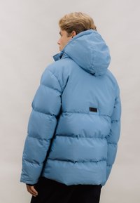 Lyseblå boblejakke med hette, som har et quiltet design og elastiske mansjetter. Et lite svart merke er synlig på baksiden.