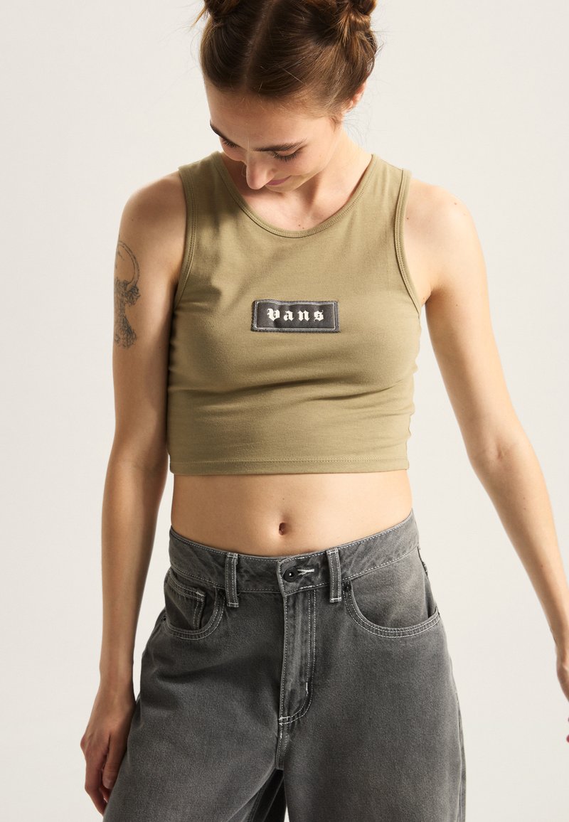 Beskuren olivgrön linne med en svart Vans logotypapplikation, i kombination med högmidjade grå denimbyxor med framfickor och kontrasterande sömmar.