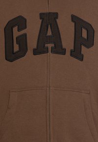 Sudadera con cremallera de color marrón, hecha de un tejido suave, con grandes letras negras "GAP" en la parte frontal y bolsillos delanteros.