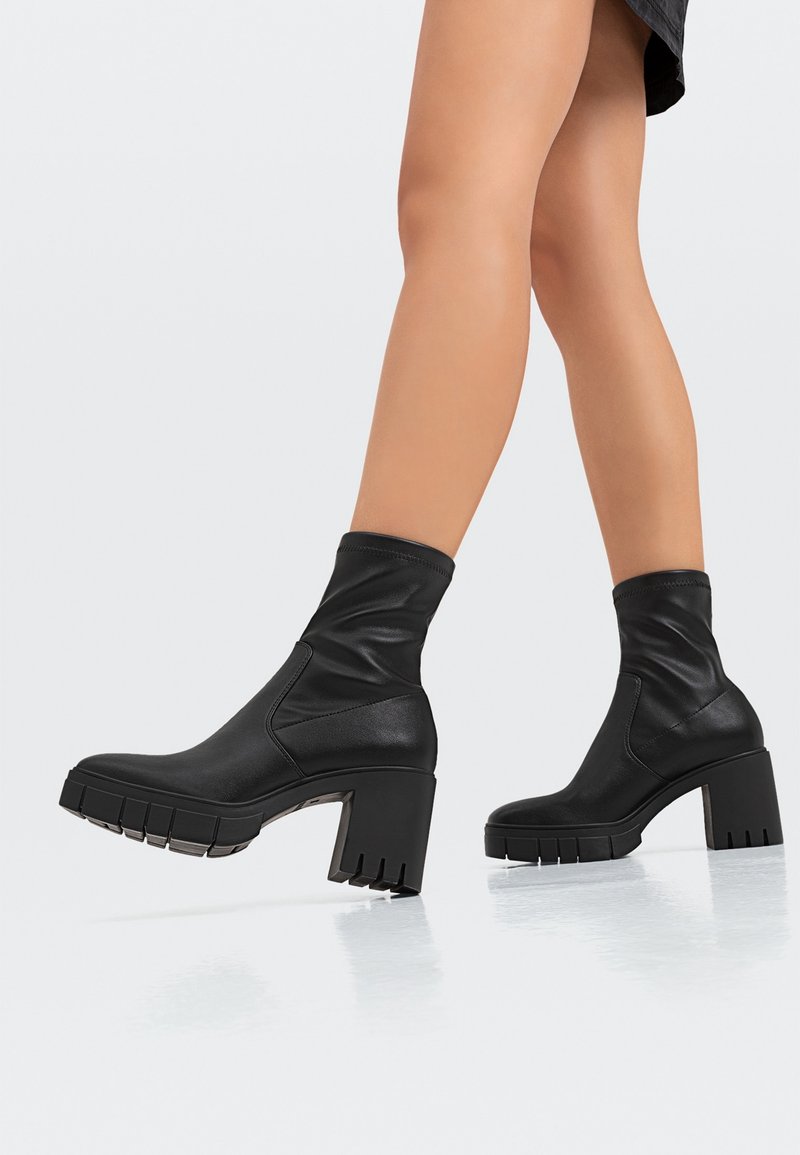 Bottines noires à talons épais portées avec des jambes nues, marchant sur une surface réfléchissante claire.