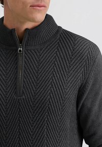 Dunkelgrauer gestrickter Pullover mit hohem Kragen, versehen mit einem Zickzack-Muster und einem Viertelreißverschluss am Hals.