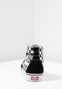 Sneakers typu high-top w czarno-białą szachownicę, zamszowy materiał, wyściełany kołnierz oraz biała gumowa podeszwa z czerwoną logo na pięcie.