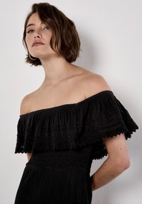 Robe noire dénudée des épaules avec un col à volants superposés, un tissu texturé et un corsage ajusté, présentant un détail de ourlet ondulé subtil.