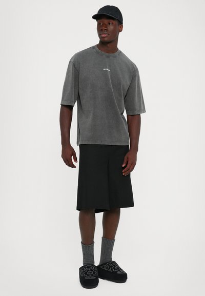 Jonge man die een vervaagde grijze oversized t-shirt, zwarte knielange shorts, grijze sokken, zwarte geborduurde slippers en een zwarte pet draagt, staande tegen een witte achtergrond.